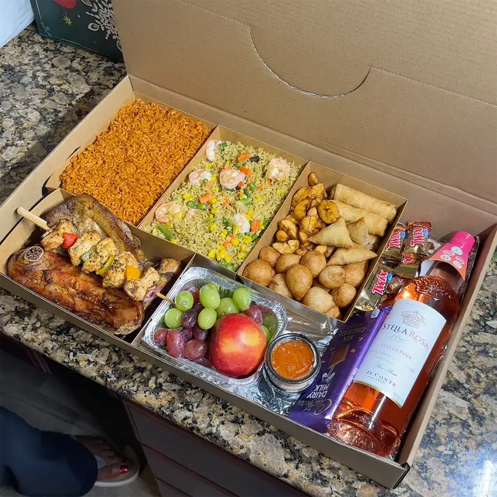 A box combo of small packs of food. Namely: jollof rice, fried rice, finger foods, peppered turkey, chicken kebab, some fruits, some chocolates, and a bottle of wine.
